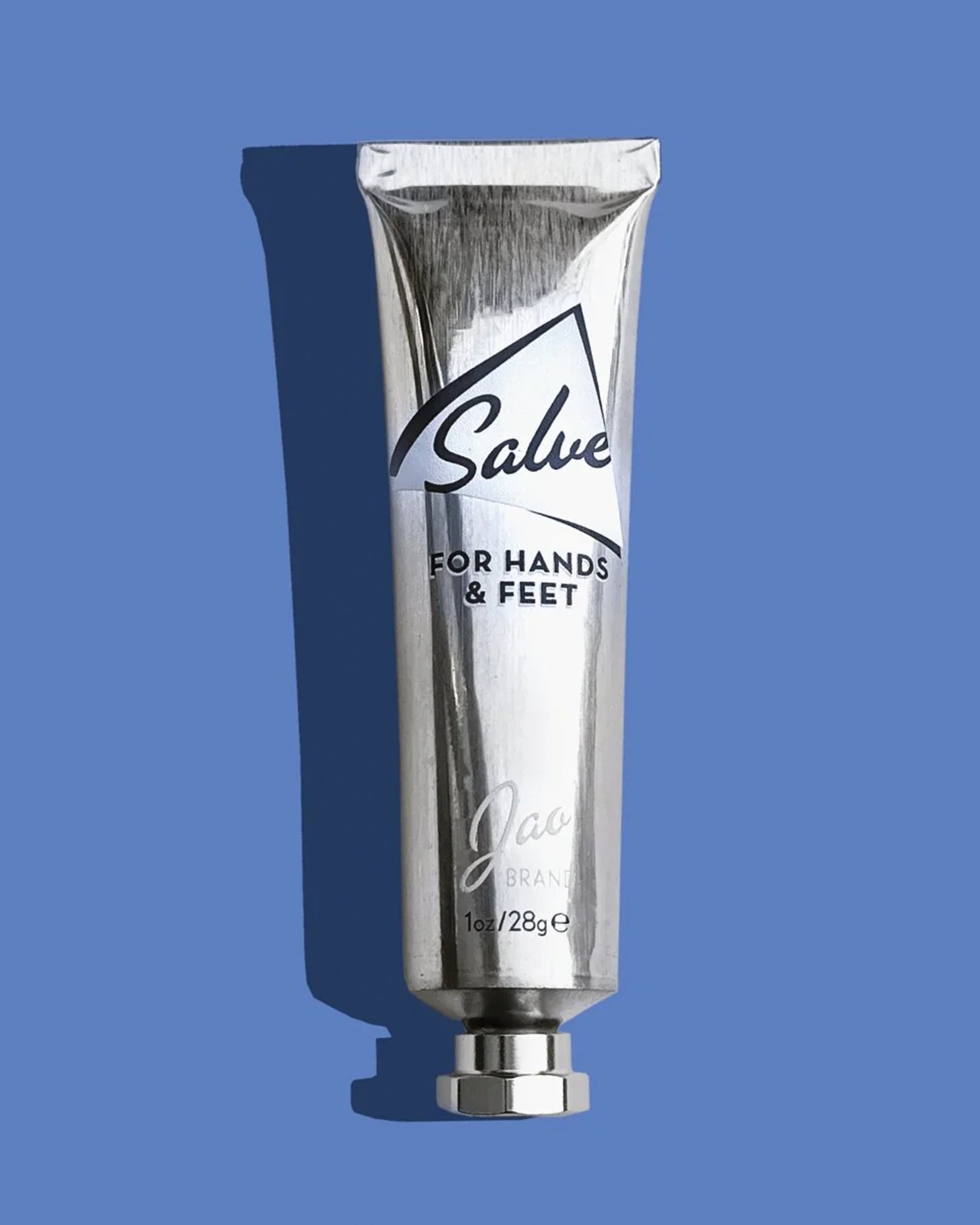 A metallic tube labeled "Jao Hand Salve, Jao Limited, 1 oz/28g" stands upright on a blue background with a left-cast shadow—ideal for moisturizing dry hands and feet with nourishing shea oil.
