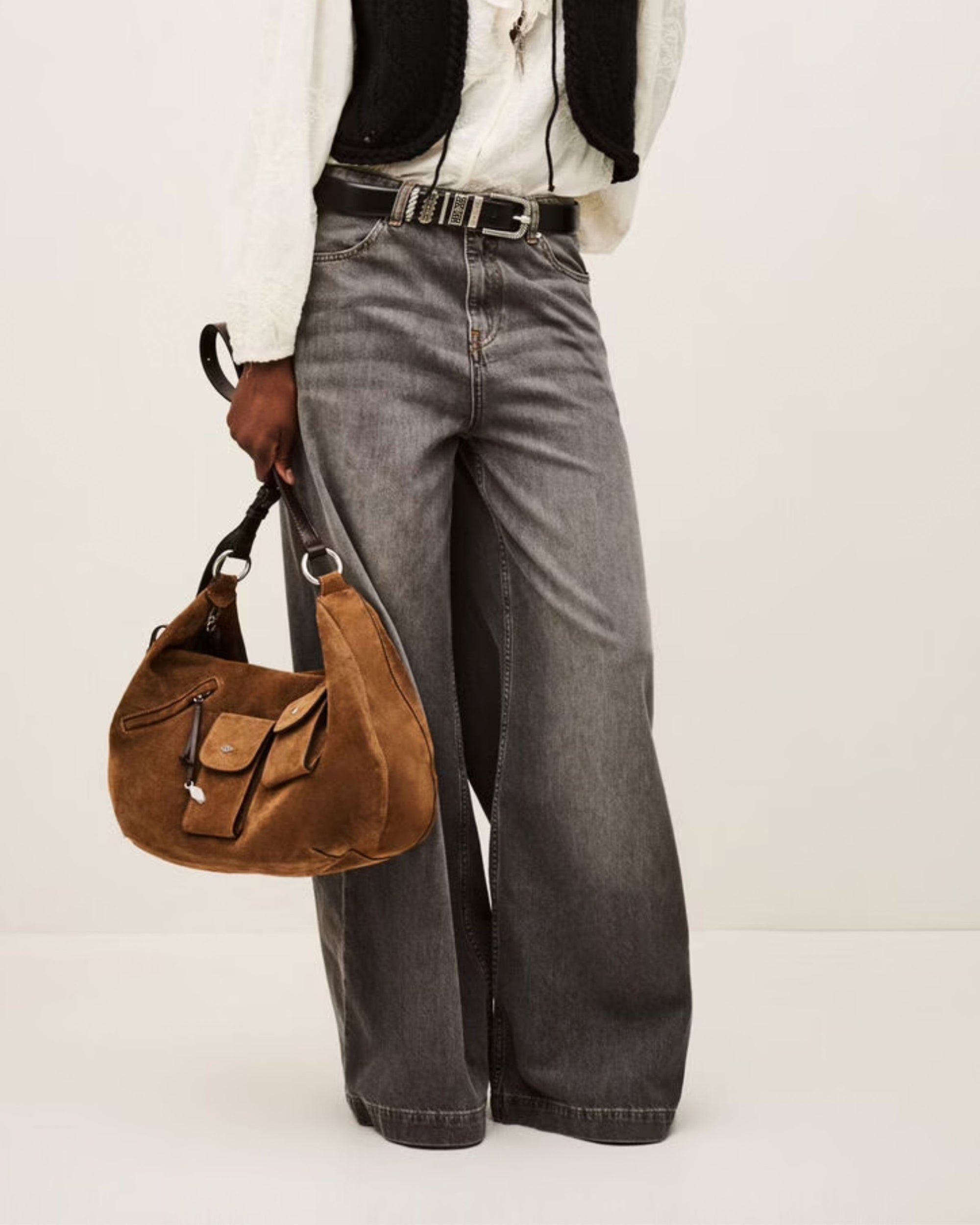 A person wearing BA&SH’s Jean Yusc—high-waisted, wide-leg gray jeans—pairs them with a white blouse and black vest, holding a brown suede shoulder bag. The image is cropped at the shoulders.