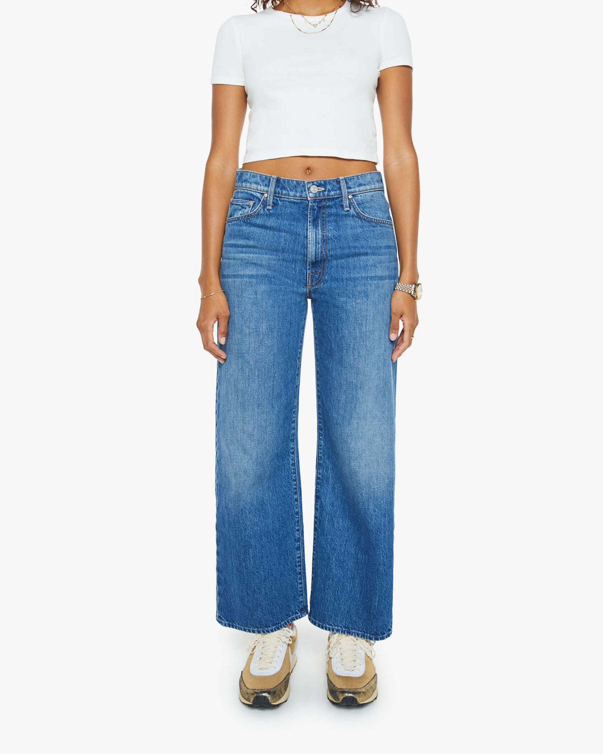 A woman wearing a white cropped t-shirt, beige sneakers, and Mother’s The Dodger Ankle straight leg blue jeans stands against a plain white background.