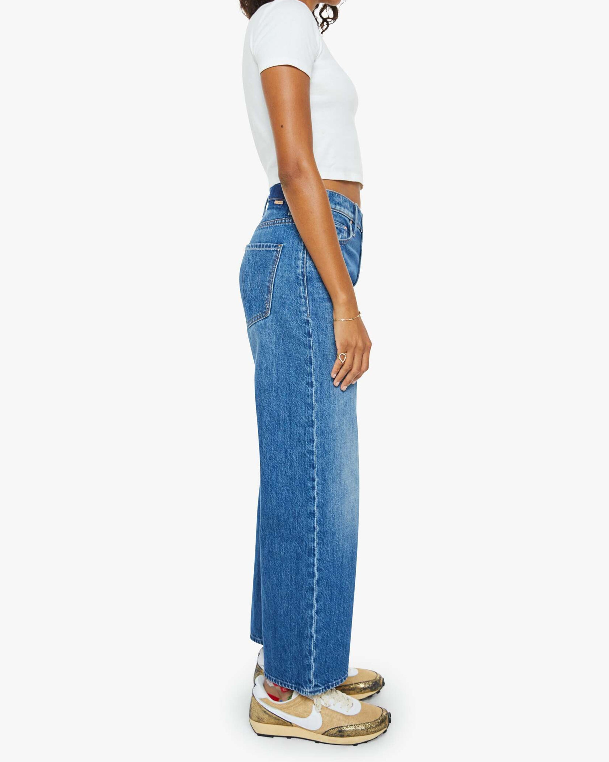 A woman with medium skin tone and curly hair stands sideways against a white background, wearing The Dodger Ankle high-waisted blue cotton blend loose straight leg pants by Mother, a white cropped tee, and worn beige Nike sneakers.