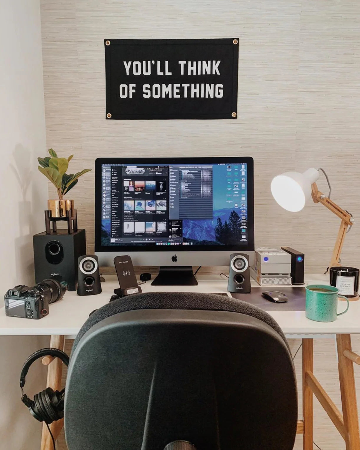 A tidy home office features an iMac, speakers, headphones, camera, plant, lamp, mug, and the Oxford Pennant "You'll Think of Something" Camp Flag 14x24—perfect for sparking inspiration and boosting creativity.
