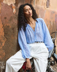 Wearing the DOEN Jonie Top and white high-waisted pants, a person with long curly hair leans against a weathered wall beside a bicycle, radiating relaxed confidence.