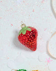 The Juicy Ornament by Accent Decor, a glass strawberry-shaped Christmas ornament (2.25" x 2.25" x 3") with a green top, rests on a sparkly surface dusted with white confetti and a light pink ribbon nearby.