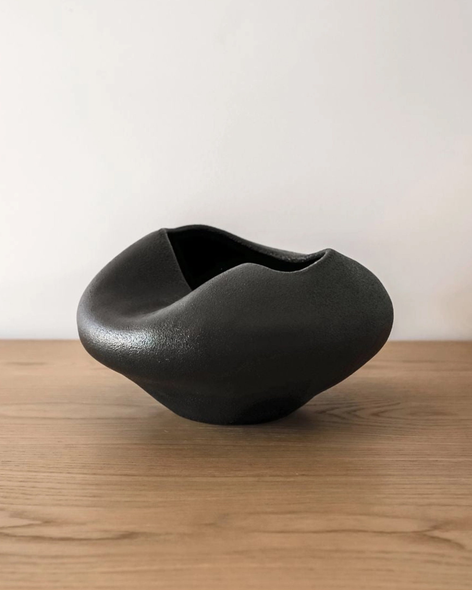 The Juliette Vase by Faire features an asymmetrical black ceramic design with a matte finish, showcasing sculptural minimalism atop a light wooden surface against a plain white background.