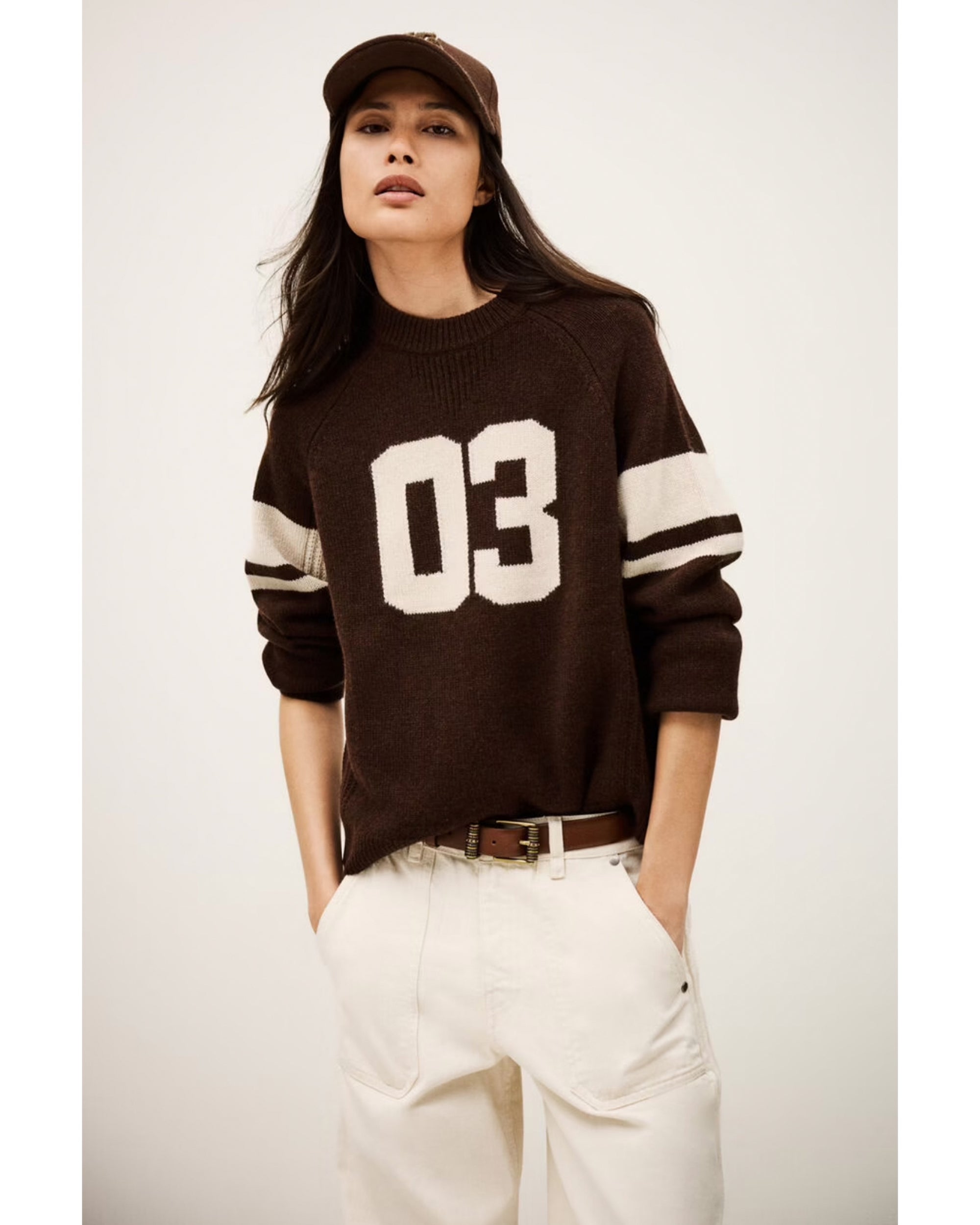 A woman wears the BA&SH Jumper Cobe, a dark brown cashmere sweater with "03" and white stripes, and a brown baseball cap. She stands confidently with hands in her white pants pockets against a plain background.