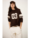 A woman wears the BA&SH Jumper Cobe, a dark brown cashmere sweater with "03" and white stripes, and a brown baseball cap. She stands confidently with hands in her white pants pockets against a plain background.