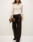 A person wears the BA&SH Jumper Banny, a beige ribbed long-sleeve top, paired with black wide-leg faux leather belted pants, layered necklaces, and a brown handbag against a plain background.