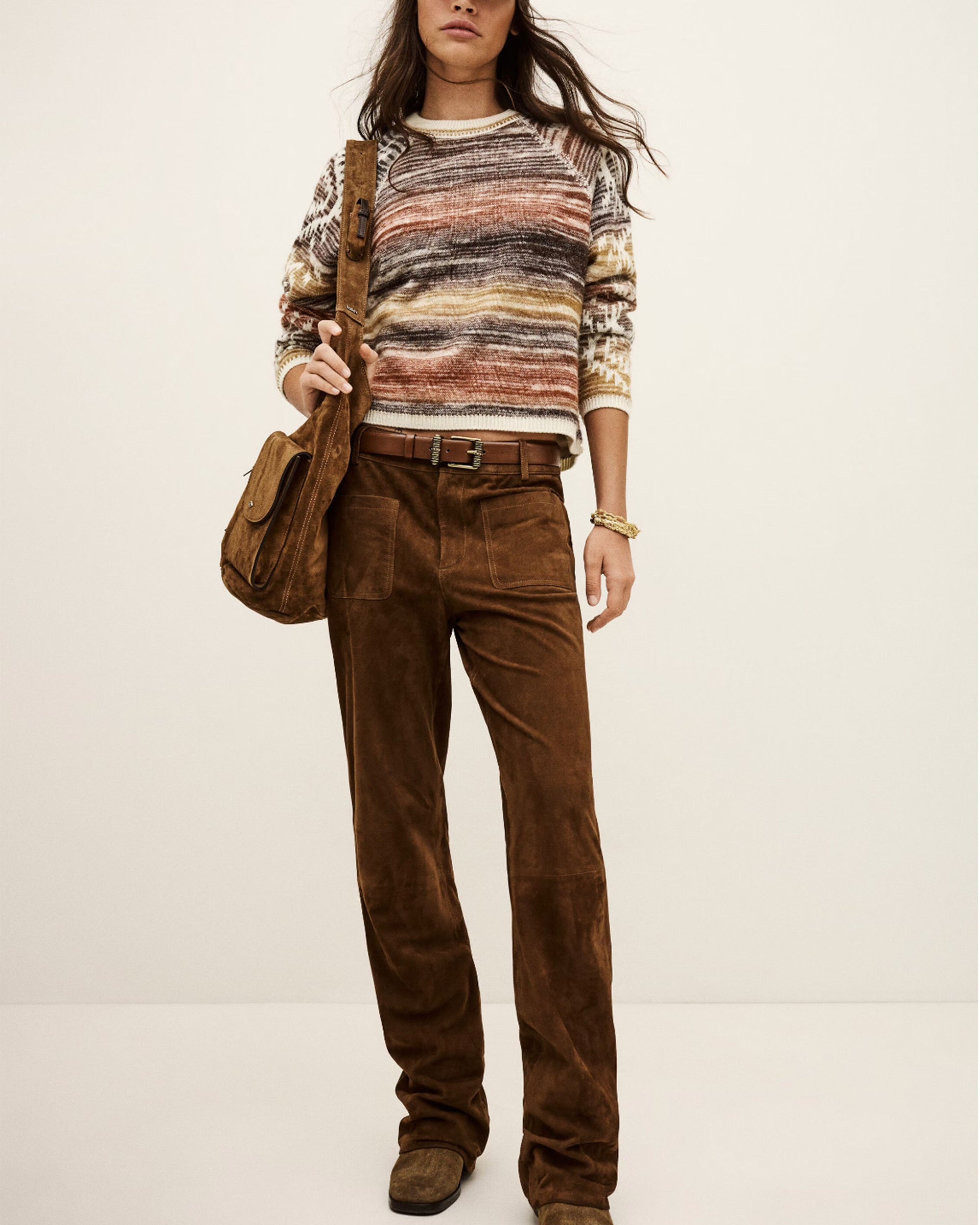 A woman wears the BA&SH Jumper Clodie— a wool sweater with brown-toned stripes—paired with brown suede pants, brown shoes, and a matching brown shoulder bag, standing against a plain light background.