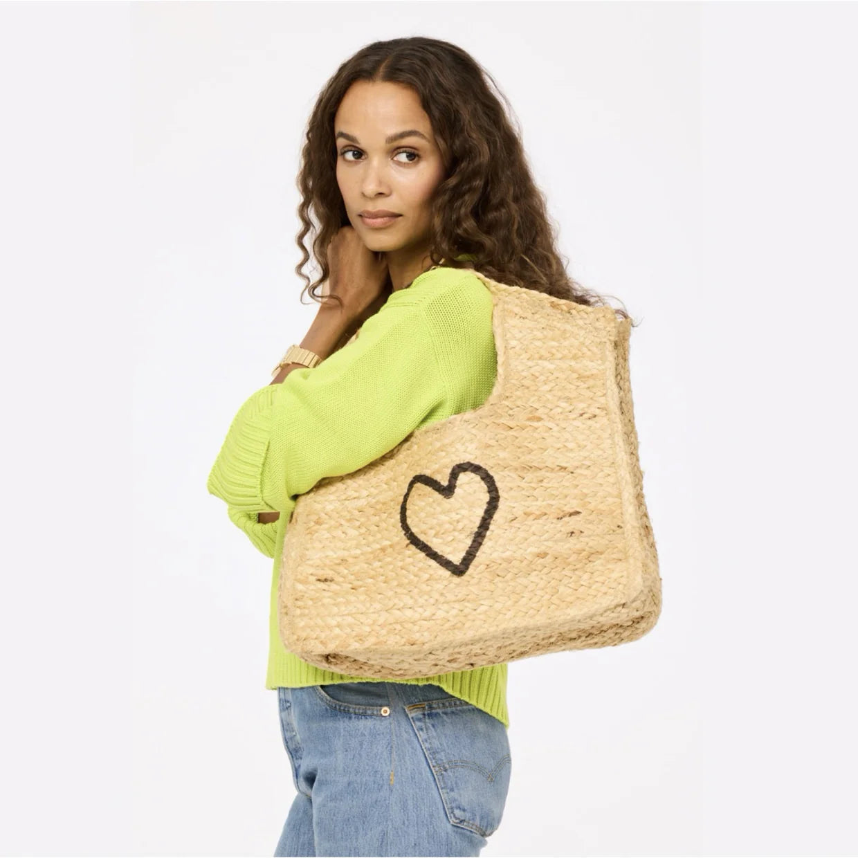 A woman with long curly hair in a light green sweater and blue jeans carries the Kerri Rosenthal Imperfect Heart Jute Tote, a large handmade bag with a black heart design, while standing against a plain light background.