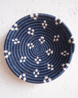 The Kabaya Small bowl by Amsha - Faire is a round Rwandan sisal basket with a dark blue and white spiral pattern and geometric accents, shown on a light textured surface.