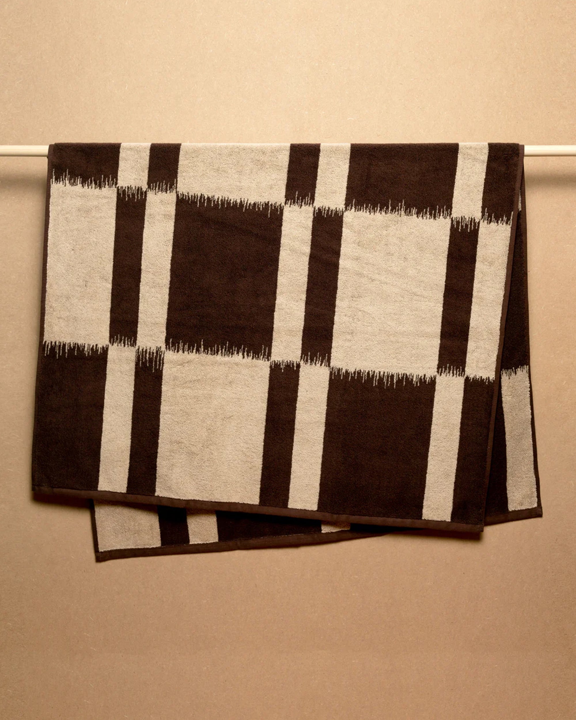 The Karin Bath Sheet by Autumn Sonata, with a large black and beige checkered pattern, is shown hanging neatly over a horizontal rod against a neutral beige wall.