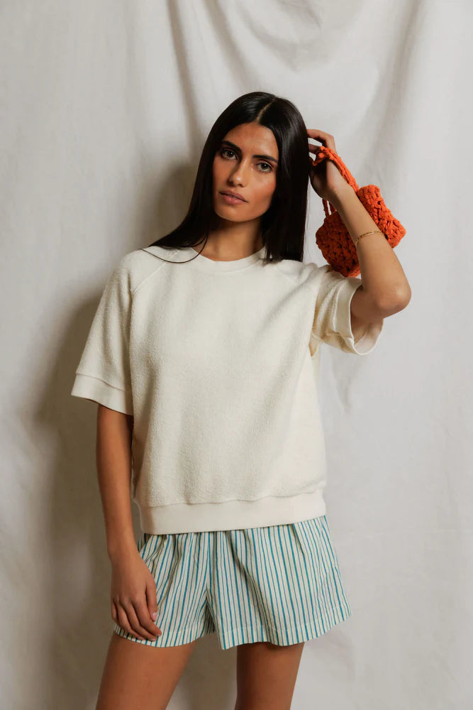 A woman with long dark hair wears the Perfectwhitetee Kennedy SS Crew Sweatshirt and blue-and-white striped shorts, holding an orange textured accessory in her right hand, standing before a light fabric backdrop.