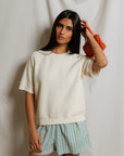 A woman with long dark hair wears the Perfectwhitetee Kennedy SS Crew Sweatshirt and blue-and-white striped shorts, holding an orange textured accessory in her right hand, standing before a light fabric backdrop.
