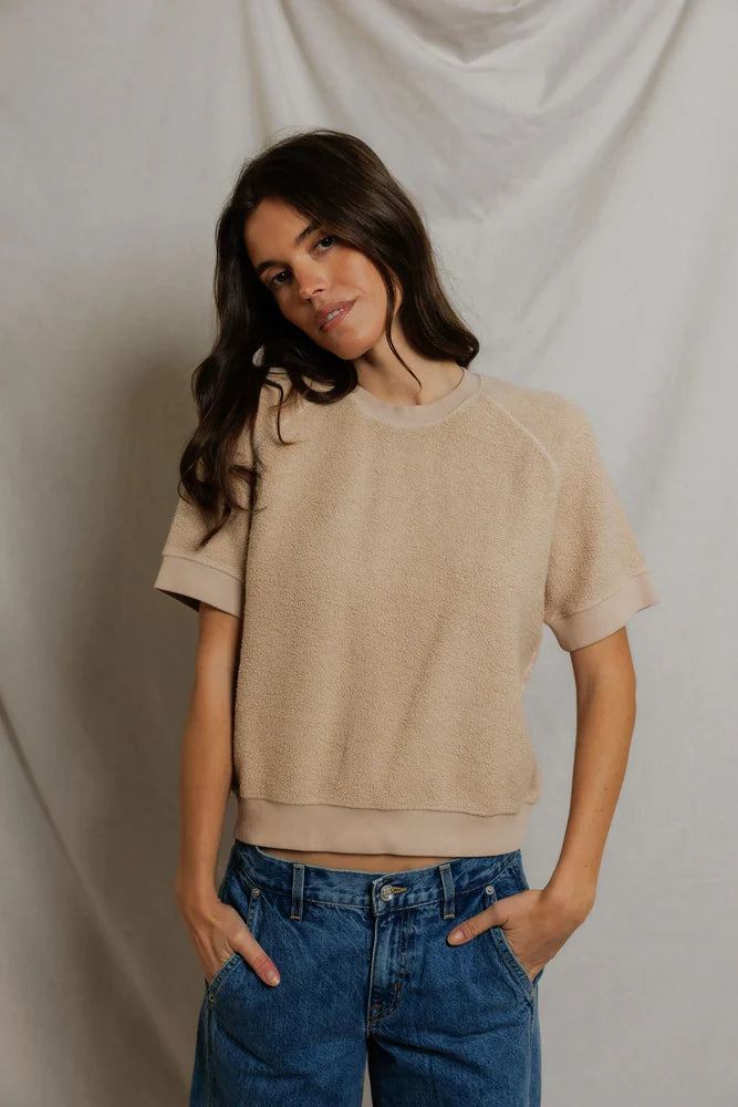 A woman with long brown hair wears the Perfectwhitetee Kennedy SS Crew Sweatshirt in beige and blue jeans, standing with her hands in her pockets against a neutral fabric backdrop.