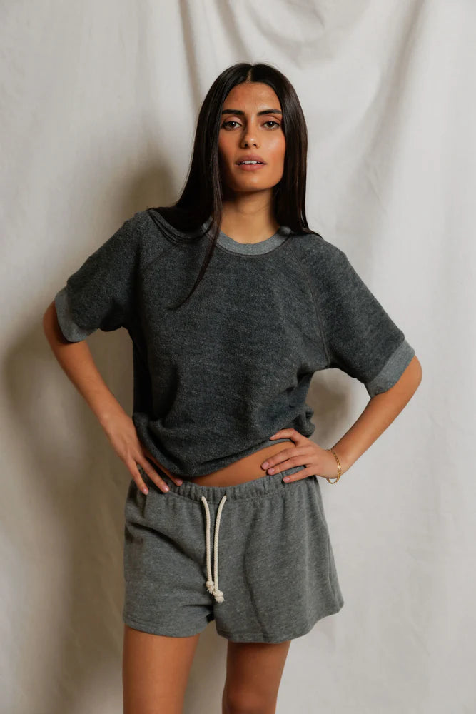 A woman with long dark hair stands before a white backdrop, wearing the Perfectwhitetee Kennedy SS Crew Sweatshirt in grey and matching drawstring shorts, one hand on her hip and a relaxed expression.