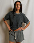 A woman with long dark hair stands before a white backdrop, wearing the Perfectwhitetee Kennedy SS Crew Sweatshirt in grey and matching drawstring shorts, one hand on her hip and a relaxed expression.
