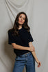 A woman with long brown hair wears the Perfectwhitetee Kennedy SS Crew Sweatshirt in a relaxed fit, paired with blue jeans. She stands before a light draped fabric background, looking at the camera with a neutral expression and one arm crossed.
