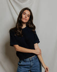 A woman with long brown hair wears the Perfectwhitetee Kennedy SS Crew Sweatshirt in a relaxed fit, paired with blue jeans. She stands before a light draped fabric background, looking at the camera with a neutral expression and one arm crossed.