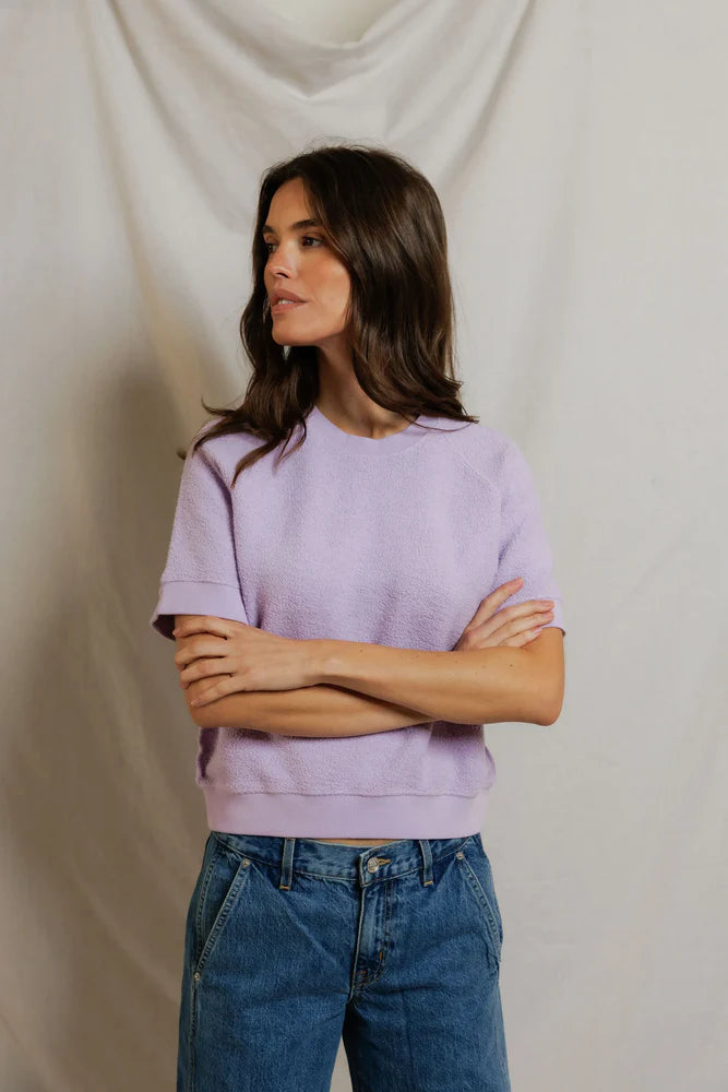 A woman with long brown hair, arms crossed, wears the Perfectwhitetee Kennedy SS Crew Sweatshirt in light purple and relaxed blue jeans, standing before a light draped fabric background.