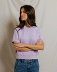 A woman with long brown hair, arms crossed, wears the Perfectwhitetee Kennedy SS Crew Sweatshirt in light purple and relaxed blue jeans, standing before a light draped fabric background.