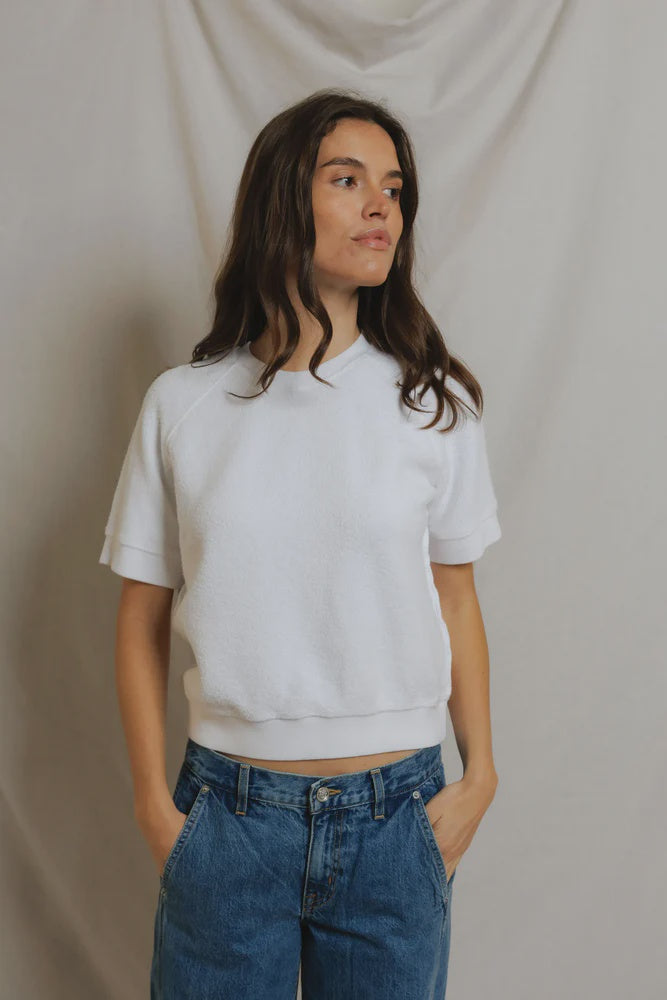 A woman with long brown hair wears the Kennedy SS Crew Sweatshirt by Perfectwhitetee and blue jeans, standing hands-in-pockets against a light draped fabric backdrop, gazing slightly to the side with a neutral expression.
