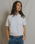 A woman with long brown hair wears the Kennedy SS Crew Sweatshirt by Perfectwhitetee and blue jeans, standing hands-in-pockets against a light draped fabric backdrop, gazing slightly to the side with a neutral expression.