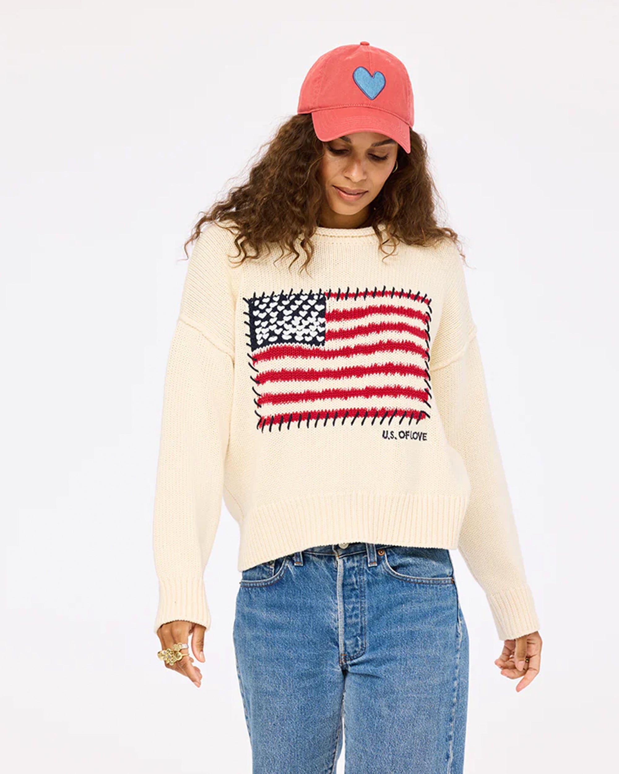 Someone with long curly hair looks downward in a neutral pose against a plain background, wearing Kerri Rosenthal’s Roll Neck /US of Love Sweater featuring a stylized American flag, blue jeans, and a red cap with a blue heart.
