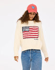 Someone with long curly hair looks downward in a neutral pose against a plain background, wearing Kerri Rosenthal’s Roll Neck /US of Love Sweater featuring a stylized American flag, blue jeans, and a red cap with a blue heart.