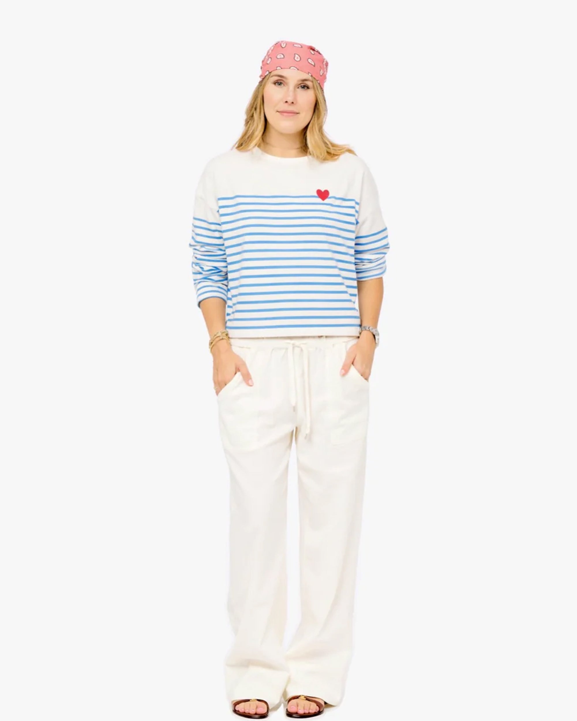 A woman faces forward, smiling slightly with her hands in her pockets. She wears a pink bandana, loose white pants, sandals, and the Kerri Rosenthal Artist Long Sleeve Stripe Tee—an oversized cotton shirt with blue stripes and a red heart on the chest.