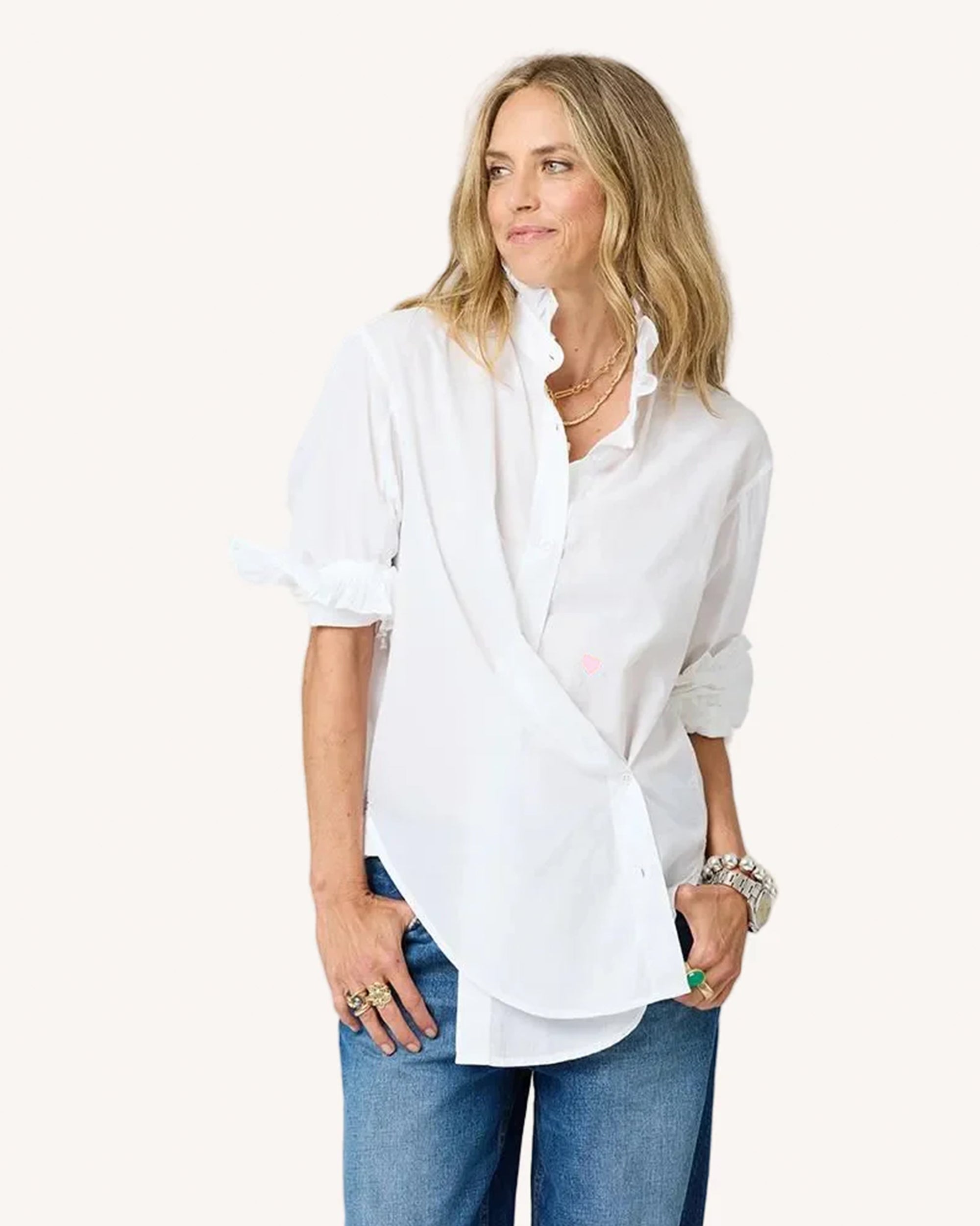 A woman with wavy blonde hair wears the Kerri Rosenthal Mia Ruffle Shirt, featuring a ruffled collar and sleeves, paired with blue jeans. She accessorizes with layered necklaces, bracelets, and rings, smiling slightly to the side.