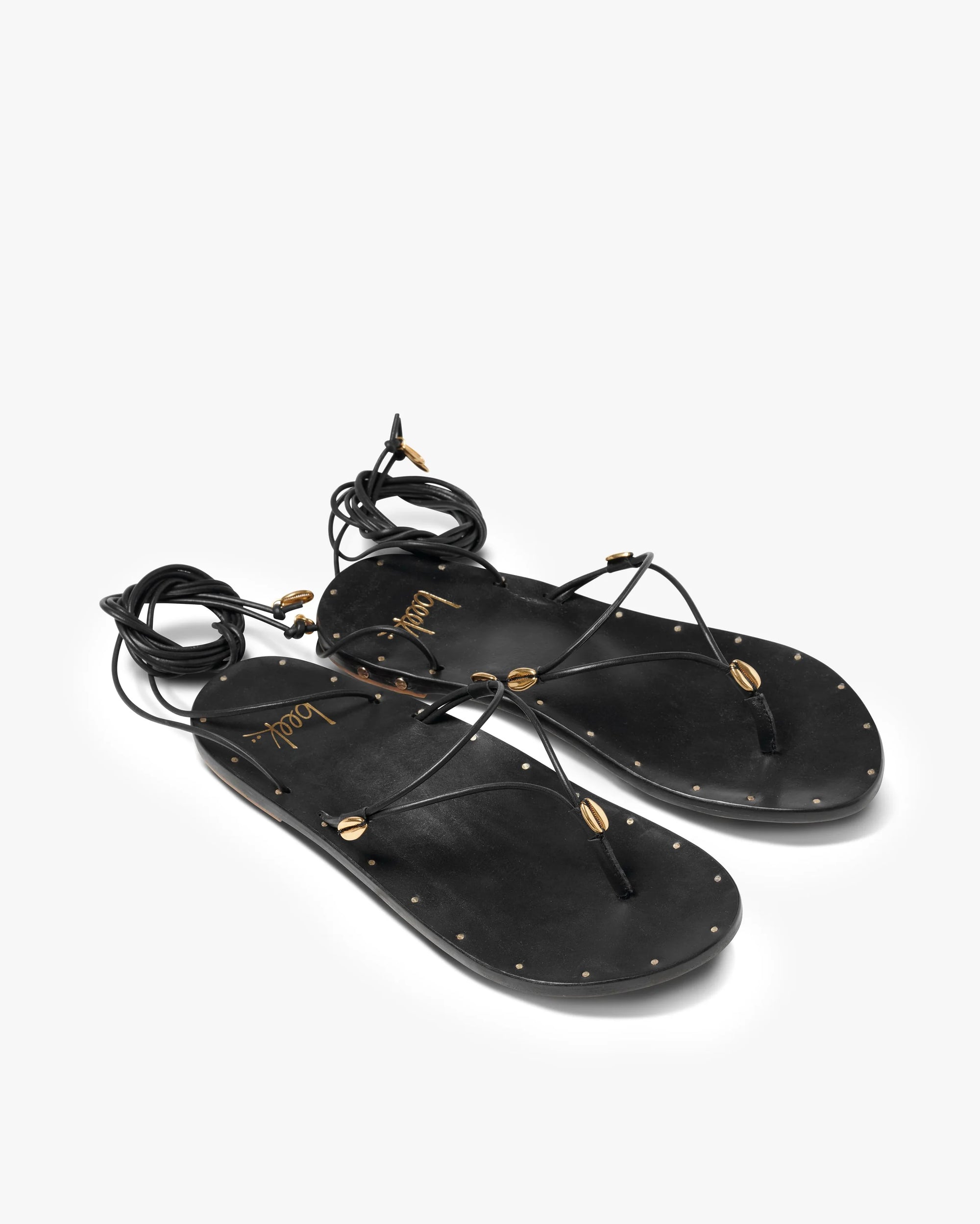 The Beek Koha Sandal features black leather, adjustable ankle-tie lace-up straps, gold bead and shell accents, and small gold studs outlining the soles, shown on a white background.