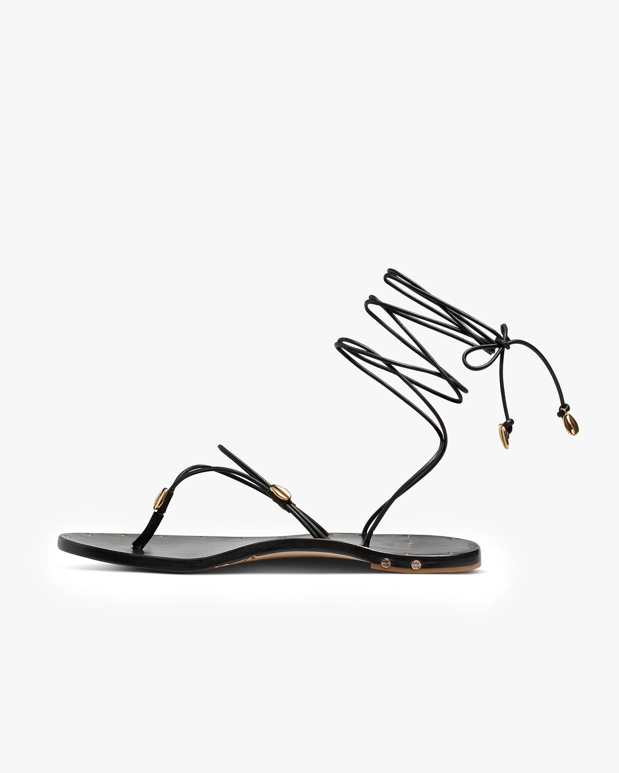 The Beek Koha Sandal is a black leather minimalist sandal with a toe strap, thin adjustable ankle lace with gold shell accents, and molded arch support for all-day comfort.