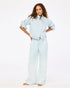 A woman with long, curly hair stands barefoot against a white background, wearing the Kerri Rosenthal Denim Jacket with rolled-up sleeves and matching wide-leg pants. She poses with hands on hips, looking straight ahead.