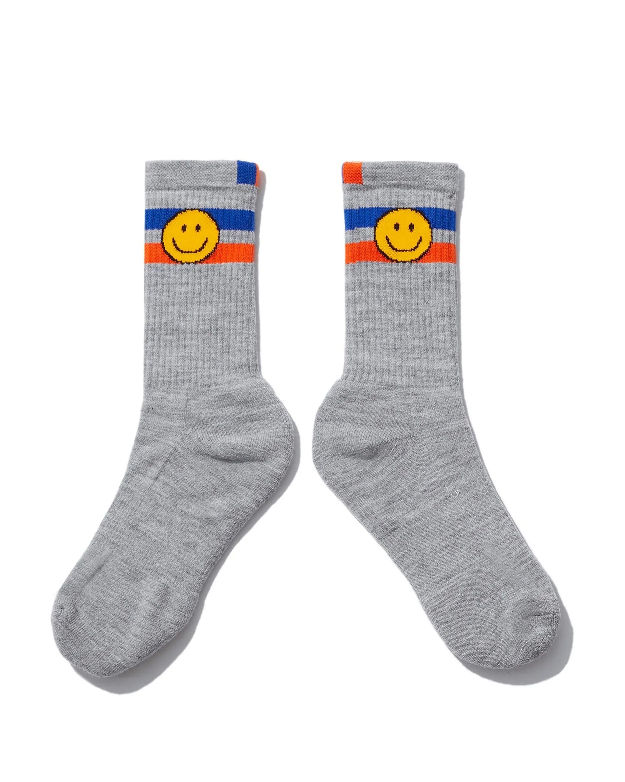 Kule's The Ribbed Smile Sock features gray ribbed fabric with blue, white, and orange stripes near the top and a yellow smiley face design below the stripes on each sock.