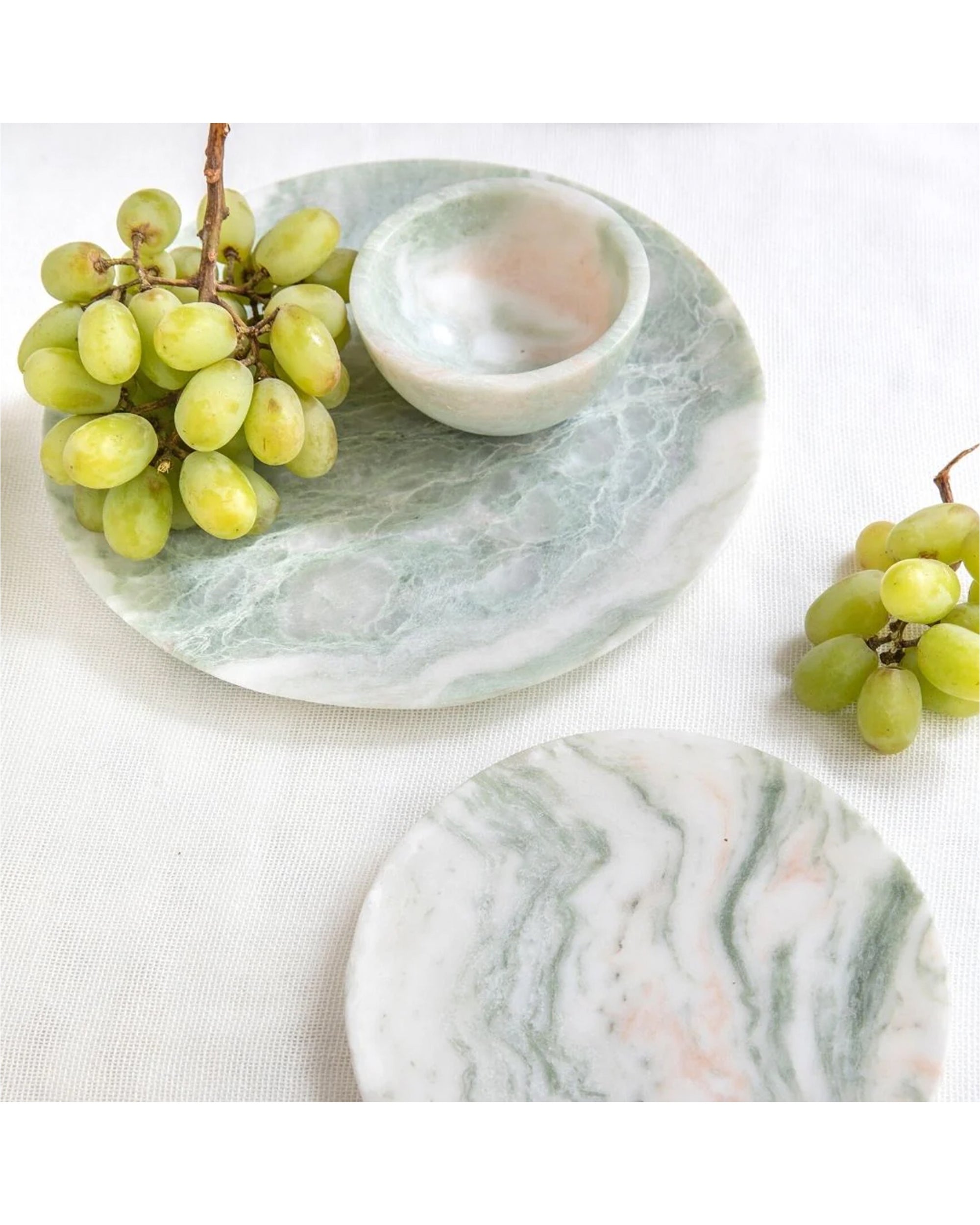 Green grapes are displayed beside and atop Be Home’s Lady Onyx Plate M, featuring green and white marble-like swirls, all arranged on a light fabric background.