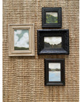 The LANDSCAPES Framed Art set by Tak Design features four small, hand-painted pieces in vintage frames arranged on a woven beige wall. Each artwork depicts clouds, hills, or water—an ideal touch of charm for any home decor.