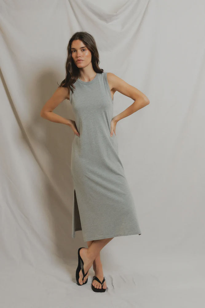 A woman with dark wavy hair stands before a light fabric backdrop wearing the Perfectwhitetee Lanell Cotton Tank Dress in light gray, a sleeveless midi with side slits, paired with black flip-flops and her arms on her hips.