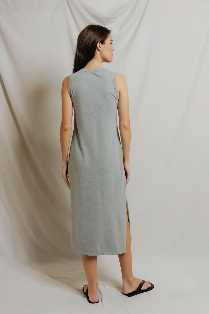 A woman with straight brown hair wears the Perfectwhitetee Lanell Cotton Tank Dress—a relaxed fit, sleeveless light gray dress with a side slit—paired with black sandals. She stands facing away, against draped, light-colored fabric.