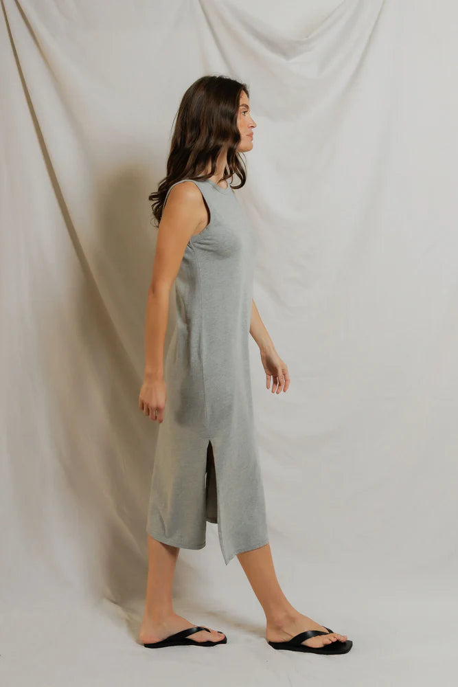 A woman with long brown hair wears the Perfectwhitetee Lanell Cotton Tank Dress, a relaxed fit gray midi tank style, along with black flip-flops as she walks in profile against a light fabric backdrop.