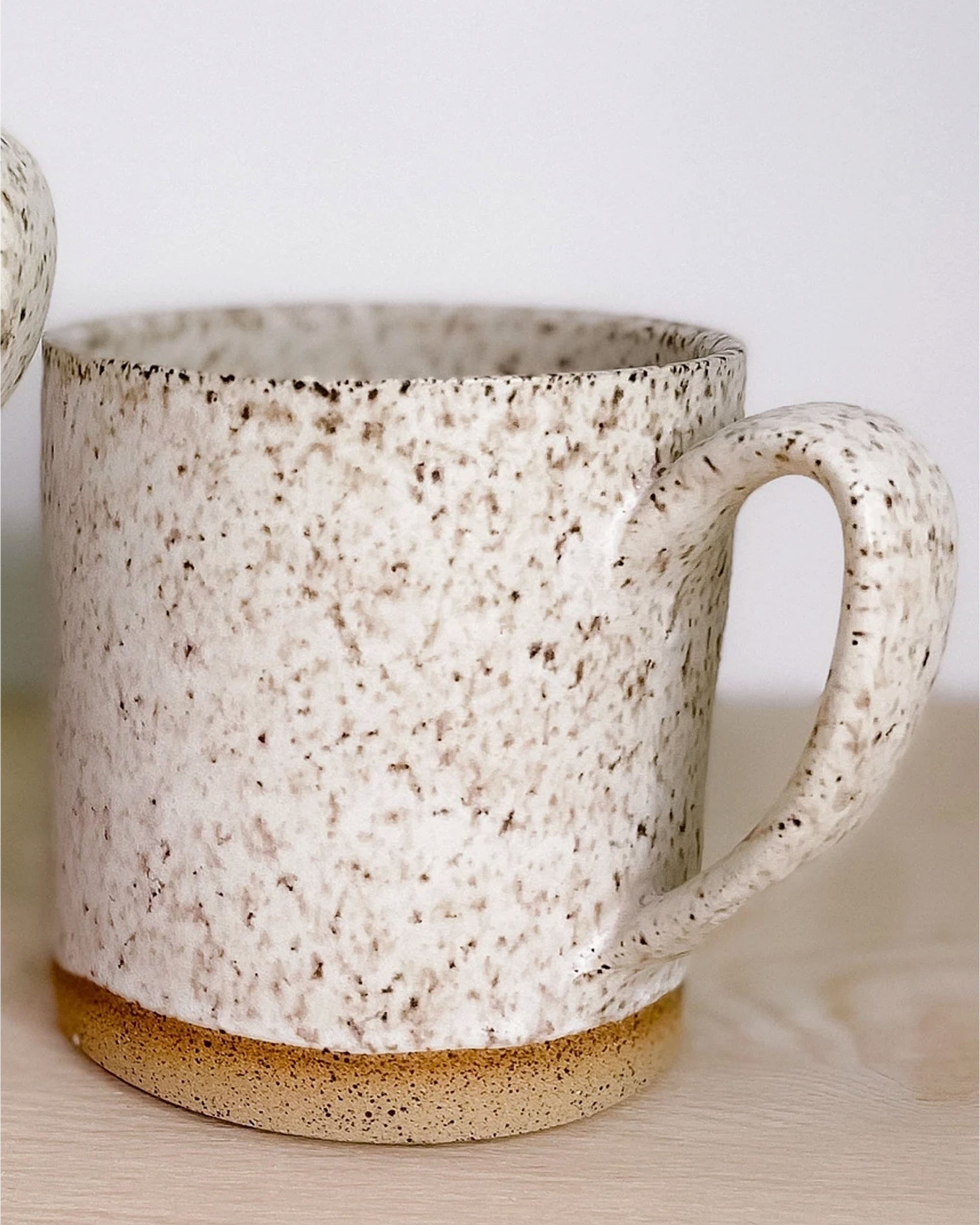 The Faire Large Speckle Mug features a hand-thrown design with a speckled white and beige glaze, rounded handle, and unglazed brown base, displayed on a light wooden surface.