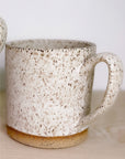 The Faire Large Speckle Mug features a hand-thrown design with a speckled white and beige glaze, rounded handle, and unglazed brown base, displayed on a light wooden surface.