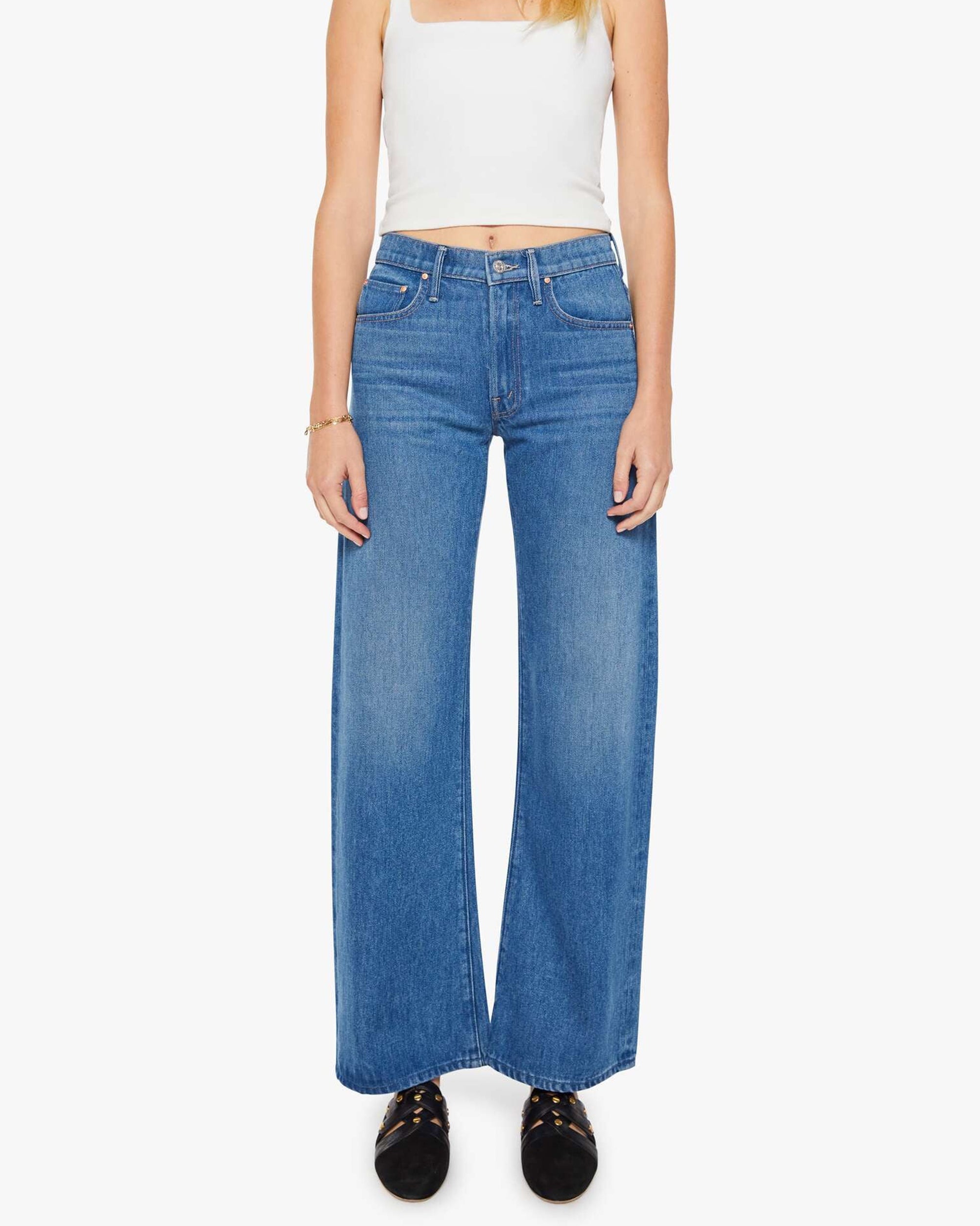 A woman stands facing forward against a white background, wearing Mother’s The Mid Rise Lasso Sneak jeans with a white sleeveless crop top and black shoes.