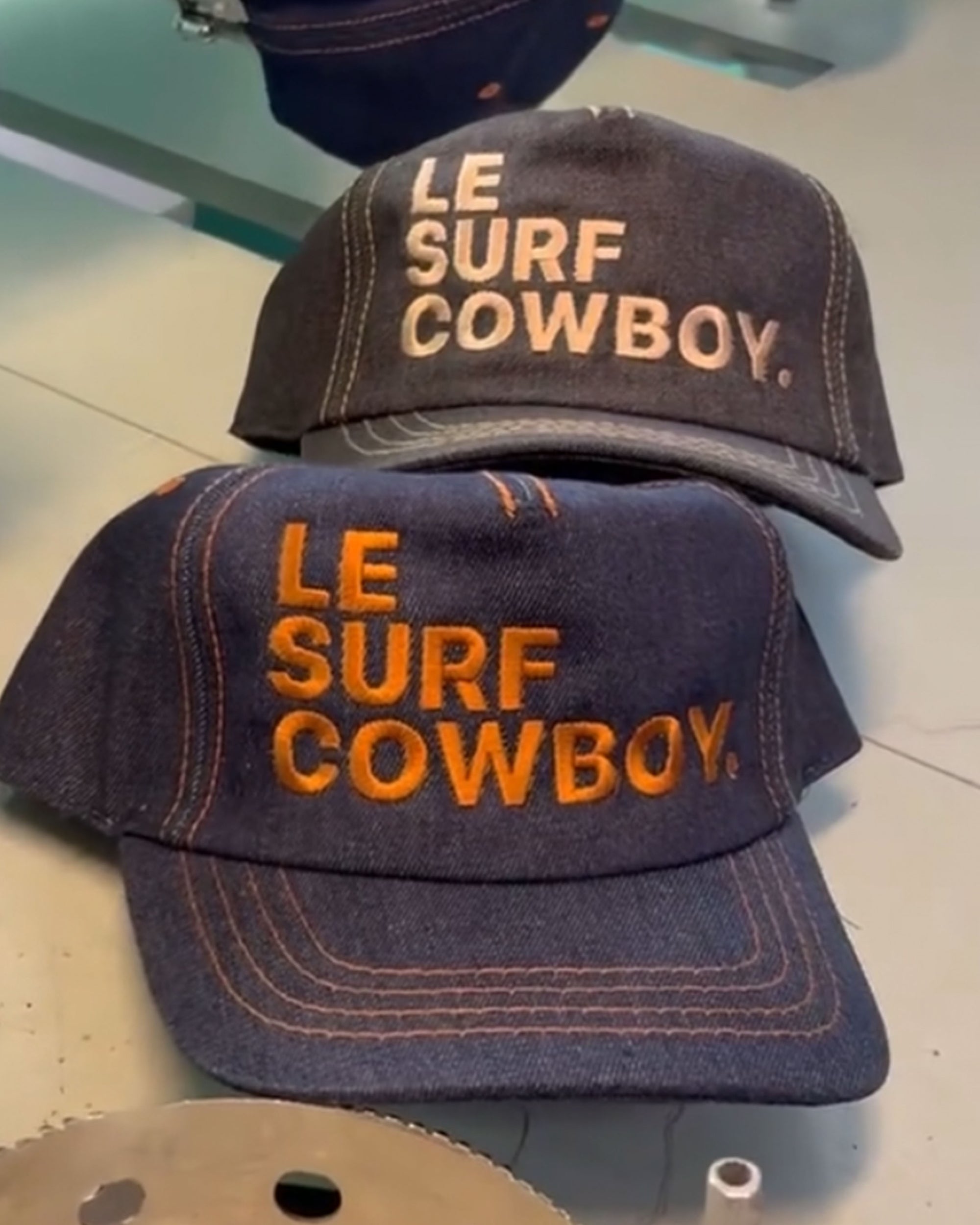 Two Le Surf Cowboy Ranch hats by Ping Pong Surf Club, featuring "LE SURF COWBOY." embroidered in white and orange thread on raw denim—an ideal tribute to classic Denim Levi style.