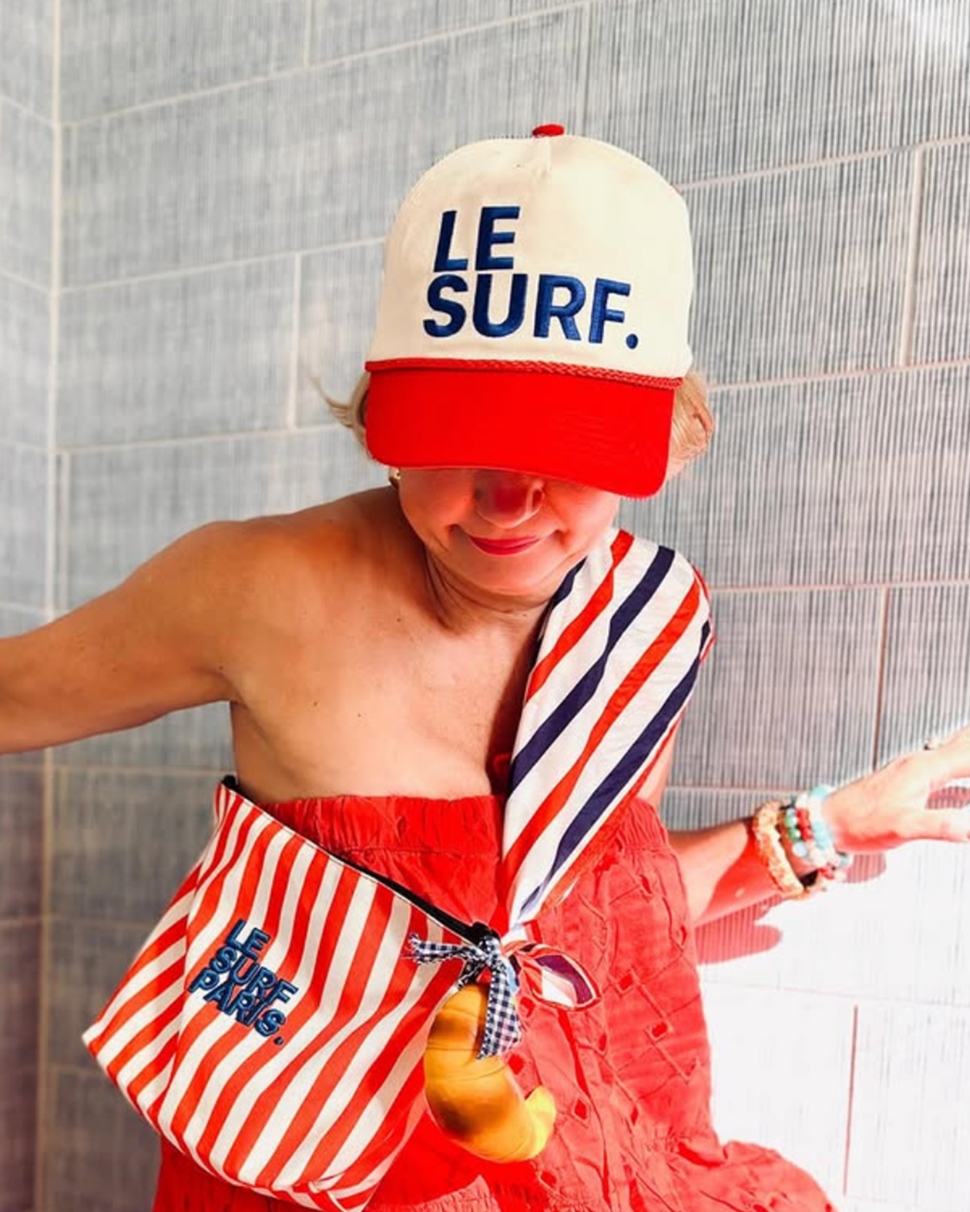 Wearing a Ping Pong Surf Club Le Surf Vintage Lesure Icon Cap, a red textured dress, striped bag and scarf, and beaded bracelets, the person poses by a tiled wall with a croissant keychain hanging from their bag.