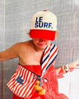 Wearing a Ping Pong Surf Club Le Surf Vintage Lesure Icon Cap, a red textured dress, striped bag and scarf, and beaded bracelets, the person poses by a tiled wall with a croissant keychain hanging from their bag.