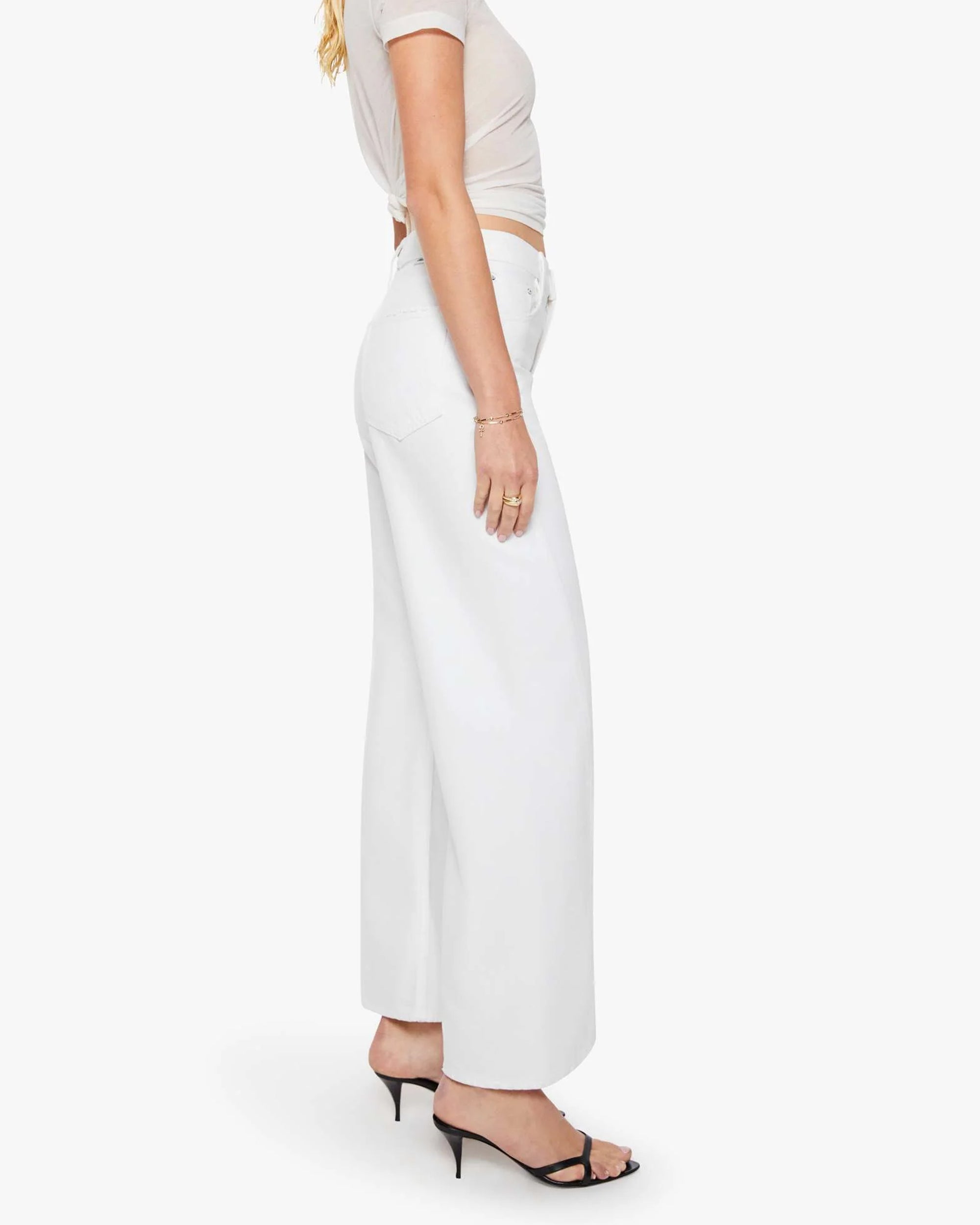 A woman stands in profile against a plain white background, wearing Mother’s The Lemon Twist Skimp high-rise wide leg pants in a rigid cotton blend, styled with a fitted white t-shirt and black heeled sandals.