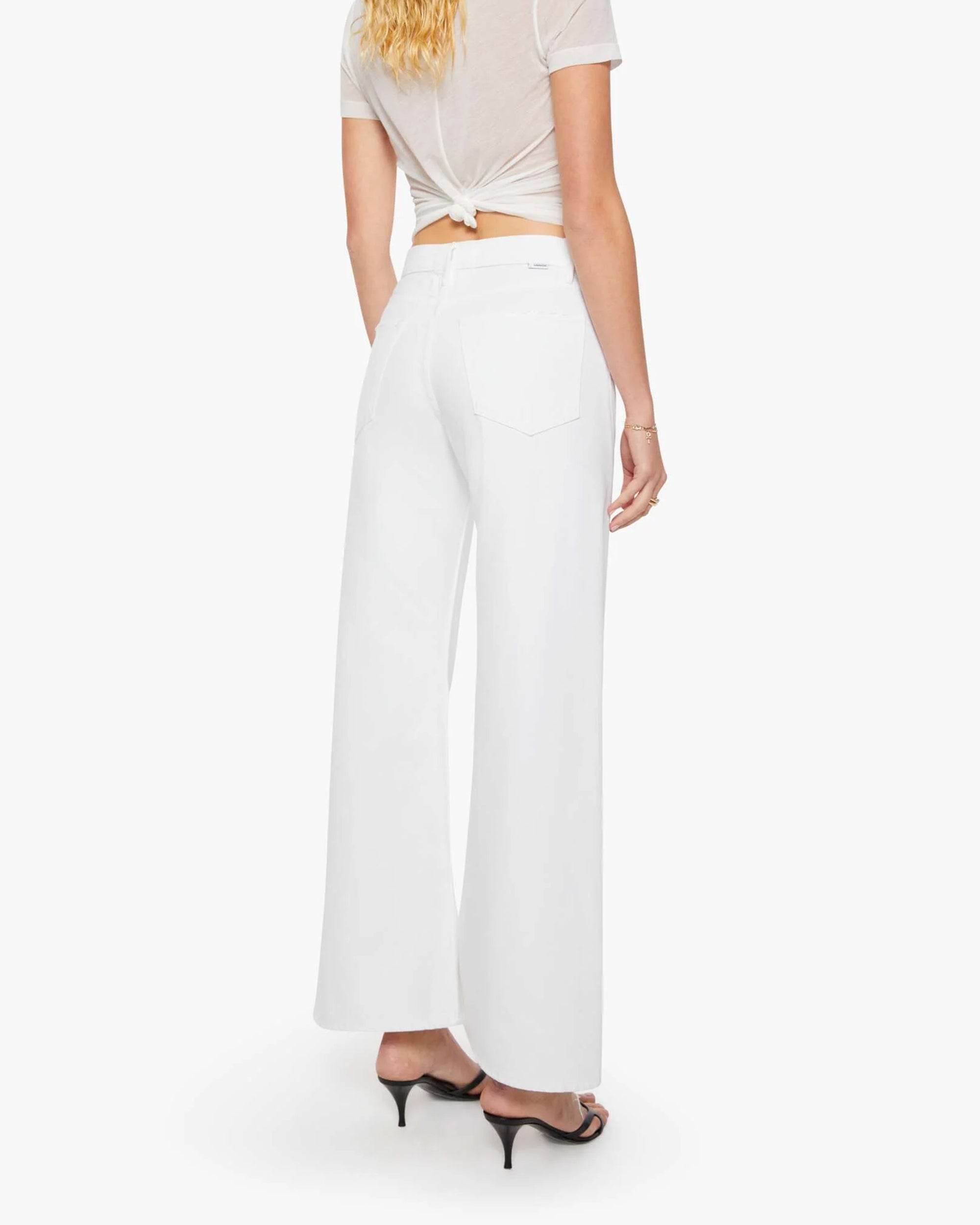 A woman faces away, wearing The Lemon Twist Skimp high-rise wide leg pants by Mother in a rigid cotton blend, paired with a sheer short-sleeve top knotted at the back and black heeled sandals. Her wavy blond hair cascades against a white background.