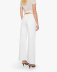 A woman faces away, wearing The Lemon Twist Skimp high-rise wide leg pants by Mother in a rigid cotton blend, paired with a sheer short-sleeve top knotted at the back and black heeled sandals. Her wavy blond hair cascades against a white background.