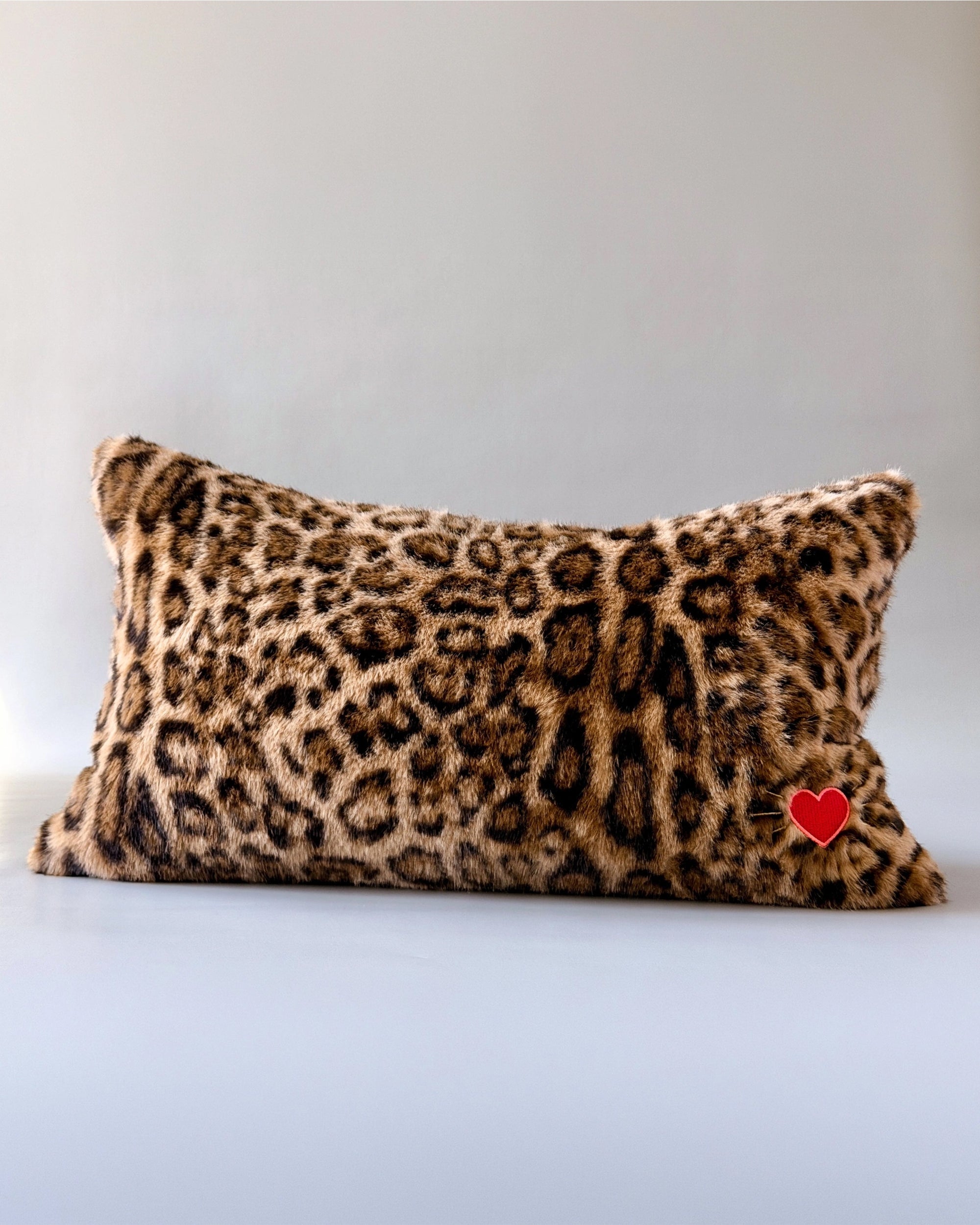 Leopard print faux fur lumbar pillow with red heart patch by Faire Muse on a light background.