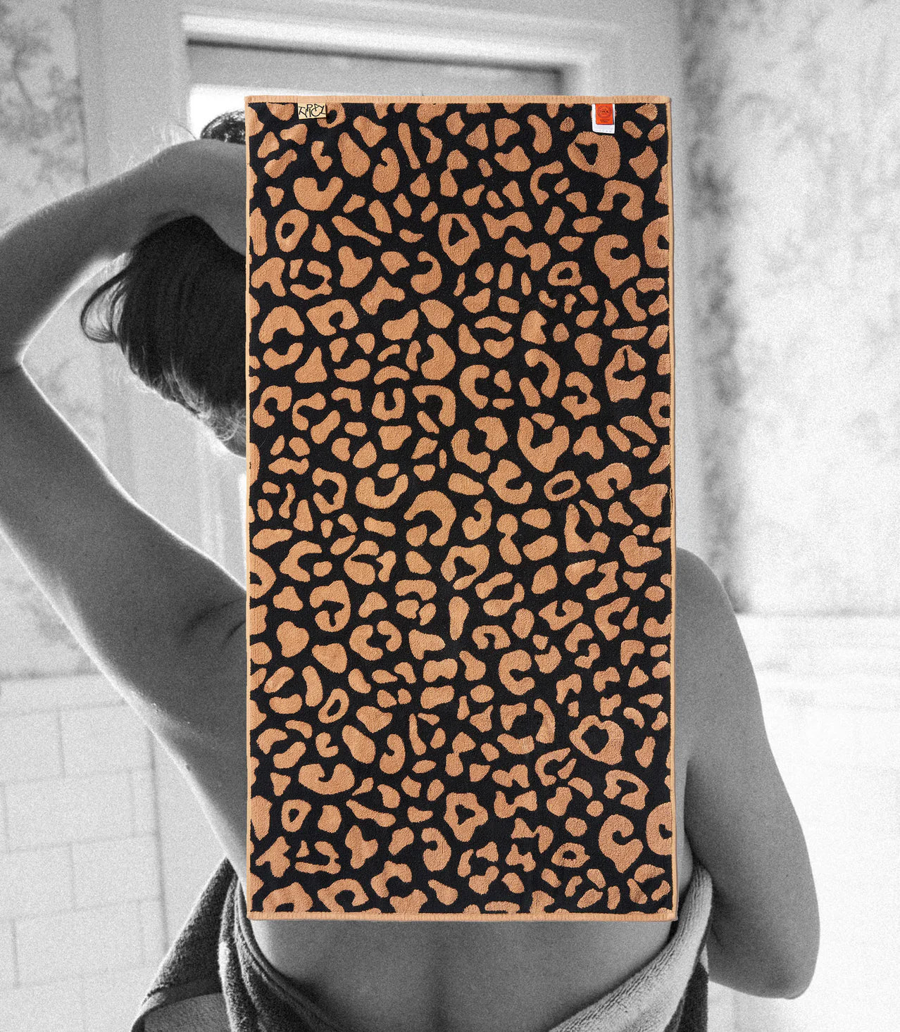 A person, back to the camera, stands in a bathroom wrapped in the Typical Wild Leopard Bath Towel. The bold leopard print towel from Typical drapes over their upper body, partially obscuring the view.